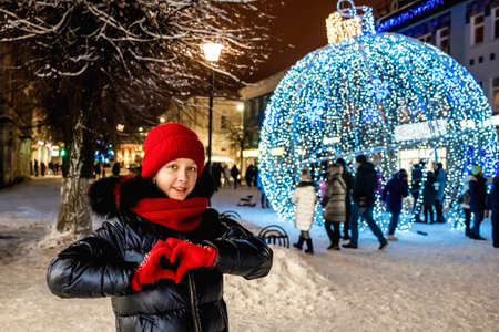 New Year's Eve. Beautiful girl with folded heart made of fingers is standing on bright festive street. Love Wishes for Christmasの写真素材