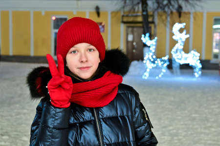pretty attractive young girl in warm clothes, wearing red hat, scarf and gloves shows victory sign "V" with her fingers against background of winter New Year decorated street. Wishes for Christmasの写真素材