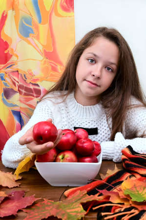 beautiful girl in knitted white sweater is sitting at table in front of plate with red apples. She holds one apple in her hand and smiles. Concept of healthy eating. Dietの写真素材