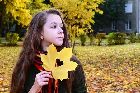 pretty girl in sweater and scarf holds yellow maple leaf with heart carved on it. Teenage girl walks...