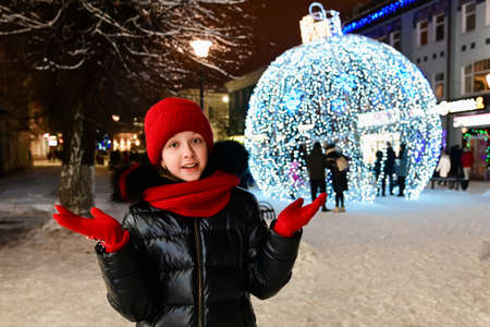 beautiful girl in warm clothes, wearing a red hat, scarf and gloves stands on New Year's decorated festive street against background of large Christmas decoration ball. New year's eveの写真素材