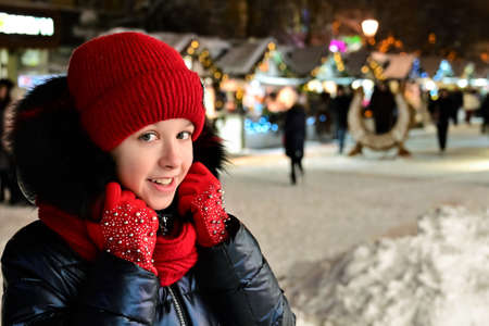 pretty smiling girl in warm clothes, wearing red hat, scarf and gloves is standing on street against...