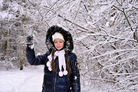 girl stands under the branches of snow-covered trees in a park. The girl is dressed in a winter...