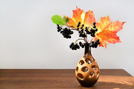 composition of chokeberry and yellow maple leaves in ceramic vase on wooden table. Autumn bouquet of black aronia berries on light background. Colorful autumn. Copy spaceの写真素材