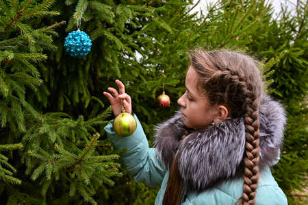 young girl with long braid and wearing warm jacket holds Christmas tree toy, ball in her hands and decorates Christmas tree outdoors. New Year's festive mood. Preparing for holidayの写真素材