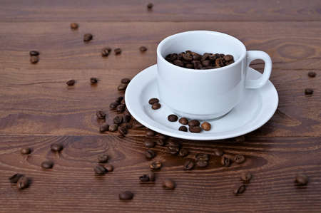 white cup filled with coffee beans on wooden table. Coffee beans lie against dark wood background....