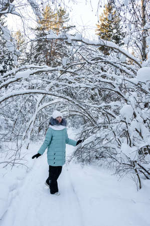 pretty stylish girl walks in winter forest among trees covered with snow. Girl is wearing warm jacket, white hat and scarf. Comfortable clothes for walking in winter. vertical photoの写真素材