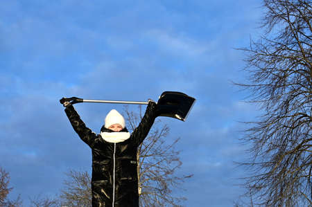young girl in winter clothes stands against background of bright blue frosty sky and holds shovel over her head. Teenage girl helps adults clean up snow and enjoy sunny dayの写真素材