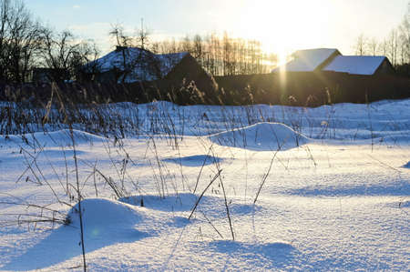 frosty winter morning in the countryside. Sun peeks out from behind roofs of houses. Everything is covered with snow, on which sun's rays slideの写真素材