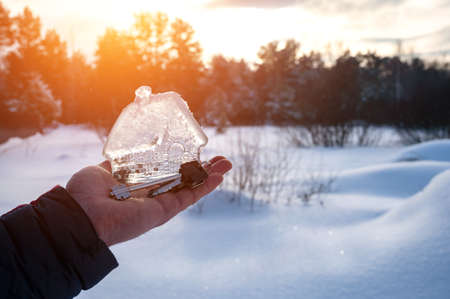 man's hand holding small glass, crystal house and keys in the palm of his hand against background of winter landscape. Artistically colored and tinted photo. Concept of dream home, take a mortgage. copy spaceの写真素材
