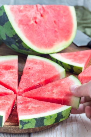 Woman hand take a slice of fresh seedless watermelon cut into triangle shape laying on a wooden plate, verticalの写真素材
