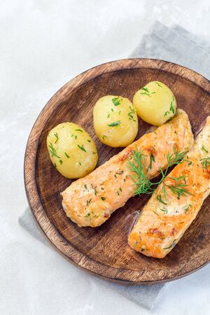 Baked salmon in creamy sauce with young boiled  potato topped with melted butter and chopped dill on a wooden plate, vertical, top viewの写真素材