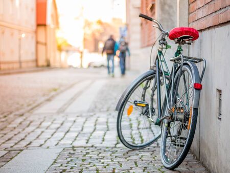 Bicycle with red seat stand near the wall on the street, sunlight and walking couple on the...