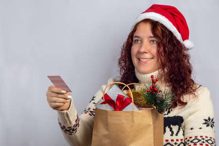 Smailing young woman in santa hat and scandinavian sweater with bag full of gifts giving credit...