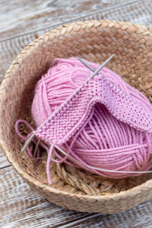 Piece of knitting with needles and pink yarn skein in a wicker basket, on a wooden background, verticalの写真素材