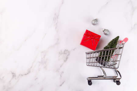Mini shopping cart with Christmas tree, red gift box and festive balls on a marble backgroundの写真素材