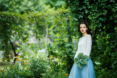 Portrait of a young slim beautiful girl with retro hair wearing a white blouse, blue skirt, posing against a background of vineyardの写真素材