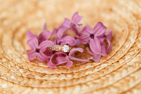 Elegant Golden engagement ring with brilliant and spring natural flowers of lilac on a straw hatの写真素材