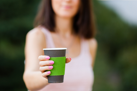 Girl is holding green paper cup, mug in hands. Mockup for products presentations, logo and text. Hand holding paper cup of coffee in a green park. Branding mockup sceneの写真素材