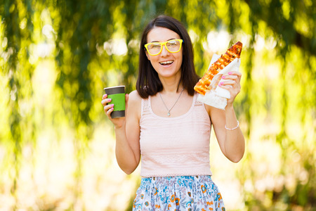 Girl is holding paper cup of coffee in hands and eating pizzaの写真素材