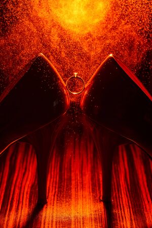 Bride's jewel ring between women's shoes with orange splashes of perfume in the orange background. Wedding concept with ring.の写真素材