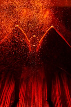 Bride's jewel ring between women's shoes with orange splashes of perfume in the orange background. Wedding concept with ring.の写真素材