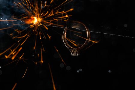 Wedding engagement ring on burning bengal light with many sparks and water spray on dark background. Marriage proposal for Christmas.の写真素材