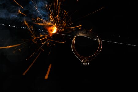 Wedding engagement ring on burning bengal light with many sparks and water spray on dark background. Marriage proposal for Christmas.の写真素材