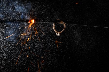 Wedding engagement ring on burning bengal light with many sparks and water spray on dark background. Marriage proposal for Christmas.の写真素材