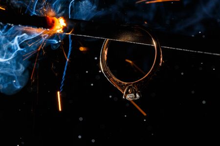 Wedding engagement ring on burning bengal light with many sparks and water spray on dark background. Marriage proposal for Christmas.の写真素材