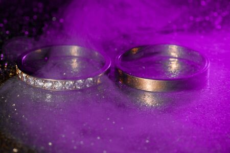 A pair of gold wedding rings with blue neon bokeh background with smoke.の写真素材