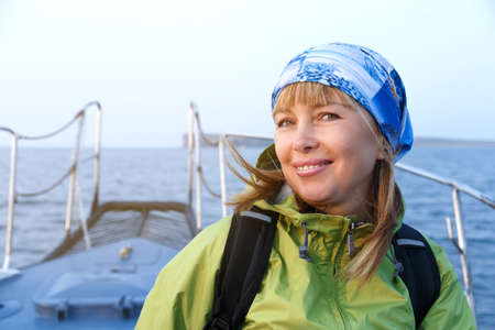 Portrait of a smiling pretty blonde woman in green jacket with backpack standing on the open deck of ship.の写真素材
