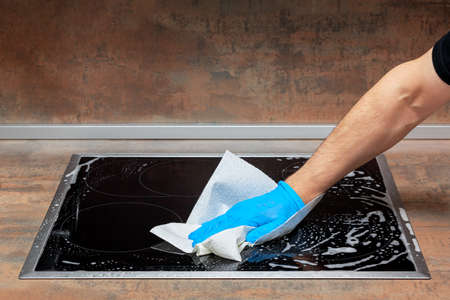 Men's hand in blue glove washes the modern electric stove with a paper napkin and cleansing foamの写真素材