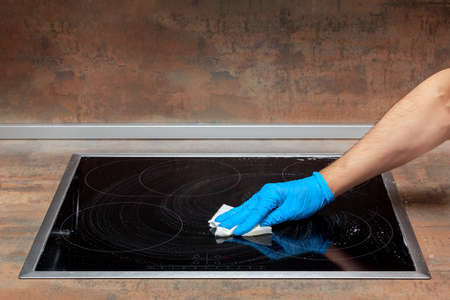 Men's hand in a blue glove removes the remaining detergent from the electric stove with a paper towelの写真素材