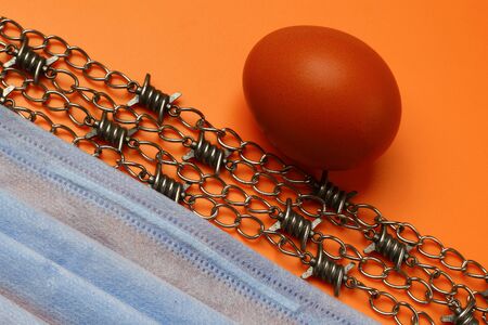 Chicken egg, barbed wire on a chain and medical mask. The concept of Easter in quarantine, closing borders, canceling flights and celebrating in isolation modeの写真素材
