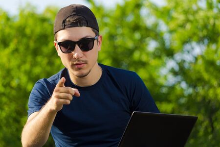 Young male student or freelancer sits in the park with a laptop and shows a finger. Concept: You can...