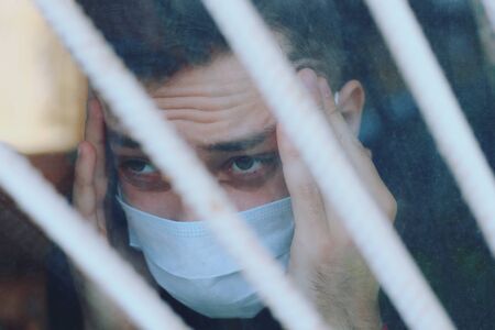 Sad young man in medical mask on quarantine. Window with bars and reflection, tonedの写真素材