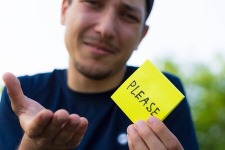 A young man holds a sticker in his hands with the inscription: PLEASE. Pitying emotion of asking for help, toned, lifestyleの写真素材