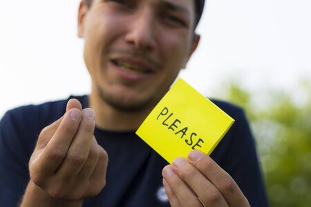 A young man holds a sticker in his hands with the inscription: PLEASE. Pitying emotion of asking for help, toned, lifestyleの写真素材