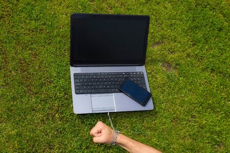 Hand tied to a laptop with a metal chain, toned. The concept of dependence on gadgets, social networks and the Internet, commitment and affection, the trap of freedom of remote work.の写真素材