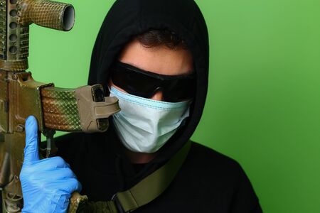 Young male gangster or criminal in a medical surgical mask and sunglasses dressed in black clothes with a hood holds an automatic assault weaponの写真素材