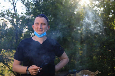 A young white man or a young adult Caucasian guy smokes a cigarette with a medical surgical mask on...