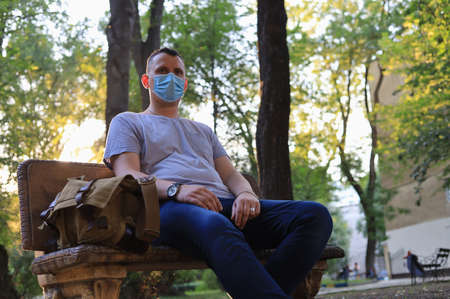 Caucasian young man or adult guy in casual clothes and a medical surgical mask is resting in a city park.の写真素材
