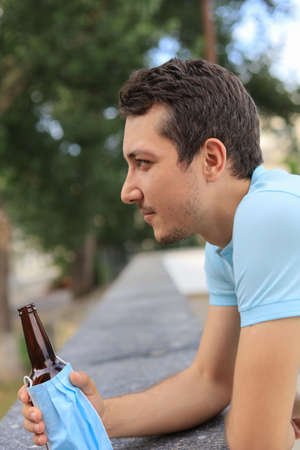 A cunning young man enjoys drinking an alcoholic drink in the city using life hack. A mask dressed on a bottle does not formally violate the law.の写真素材