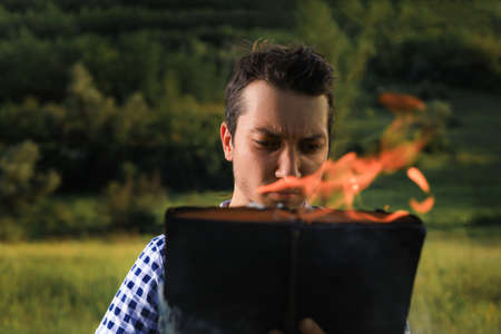 White Caucasian young man or adult guy reading a burning book on blurred forest background. Emotion...
