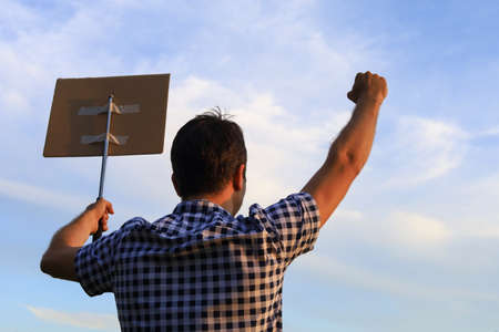 A white Caucasian young man or adult guy with a raised hand and clenched fist came out to protest. A cardboard plate is attached to a metal periscope baton. Selective focus. Blurred evening skyの写真素材