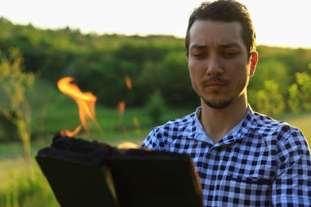 White Caucasian young man or adult guy reading a burning book on blurred forest background.