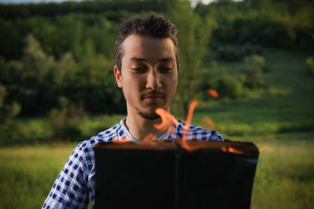 White Caucasian young man or adult guy reading a burning book on blurred forest background.の写真素材