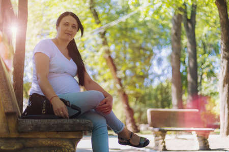 A young pregnant woman in light clothes is resting in the shade of the park's trees on a hot sunny day. Toned background. Selective focusの写真素材