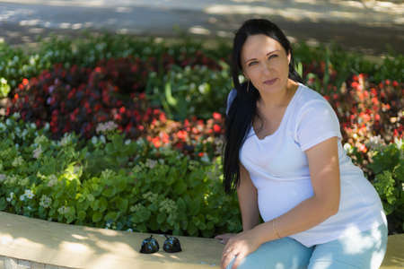 A young pregnant woman in light clothes is resting in the shade of the park's trees on a hot sunny...